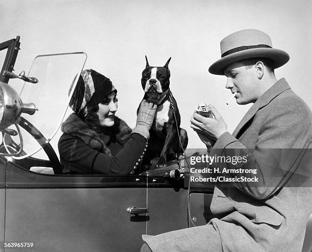 1920s WOMAN SITTING IN DRIVER'S SEAT CONVERTIBLE CAR WITH BOXER DOG MAN LIGHTING CIGARETTE