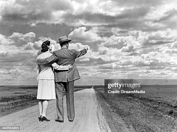 1940s BACK VIEW COUPLE WITH ARMS AROUND EACH OTHER STANDING ON OPEN ROAD POINTING TO HORIZON