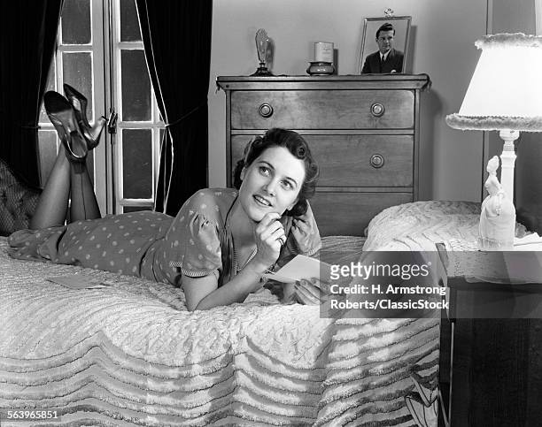 1940s FULLY DRESSED SMILING WOMAN LAYING ON HER BED WITH HER LEGS UP READING A LETTER IN HER BEDROOM