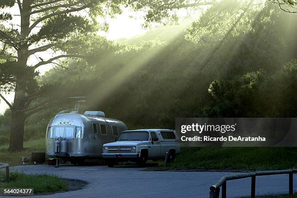 Early morning light beaming through the trees, illuminating campsite with a classic Airstream trailer at Montana De Oro State Park campground, also...