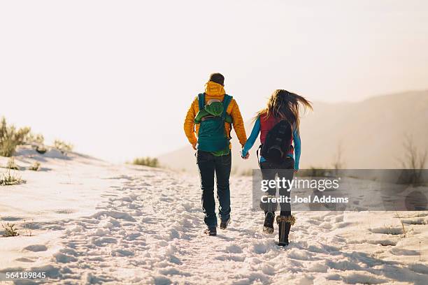 a young couple holds hands and hikes against a winter mountain scene and bright sunset. - holding hands in the snow stock pictures, royalty-free photos & images