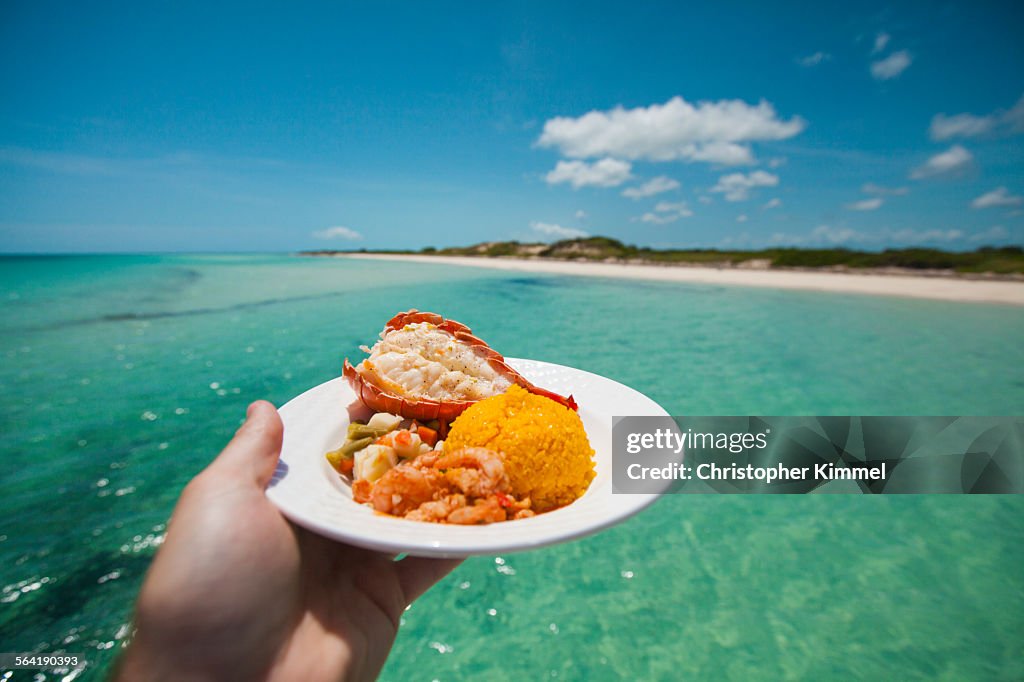 A man holds a plate of seafood with shrimp, Lobster tail, and seasoned rice while on a boat