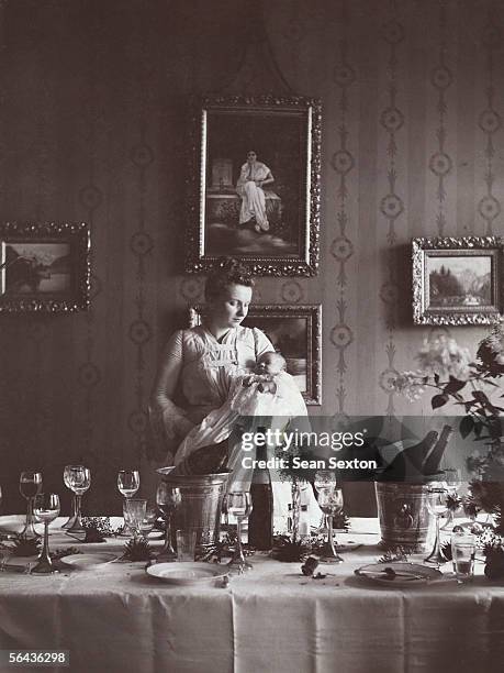 Young woman stands holding a baby next to an array of empty bottles and half finished glasses of wine, circa 1890.