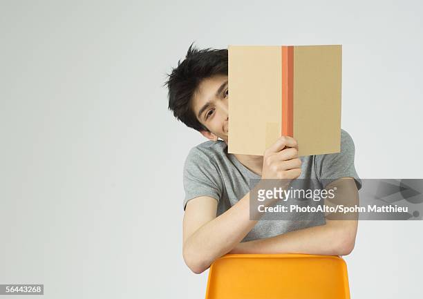 young man holding up book, peeking around the edge of it - man holding book fotografías e imágenes de stock