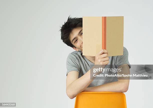 young man holding up book, peeking around the edge of it - holding book stock pictures, royalty-free photos & images