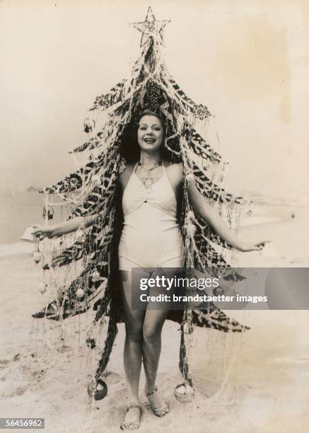 Beauty queen as Christmas tree: Girl in her bathing suit with Christmas tree dressing on a Californian beach. Photography, around 1930....