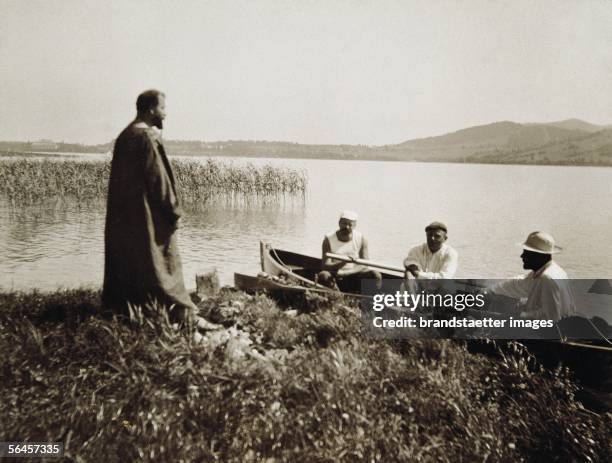 Gustav Klimt in his pinafore standing at the shore, three of his friends sitting in a rowboat at the Attersee lake. Photography, around 1910. [Gustav...