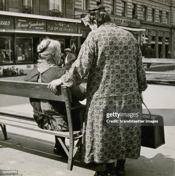 Park bench in Vienna V : old woman in flower dress with shopping bag is standing next to a park bench, another woman is sitting next to her....