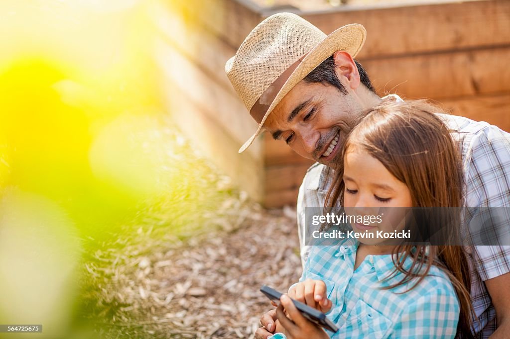 Girl on fathers lap using smartphone in community garden