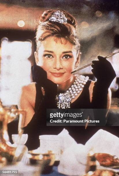 Belgian-born actress Audrey Hepburn , in a black, shoulderless dress, matching gloves, and a tiara, smiles with a cigarette holder in her hand, in...