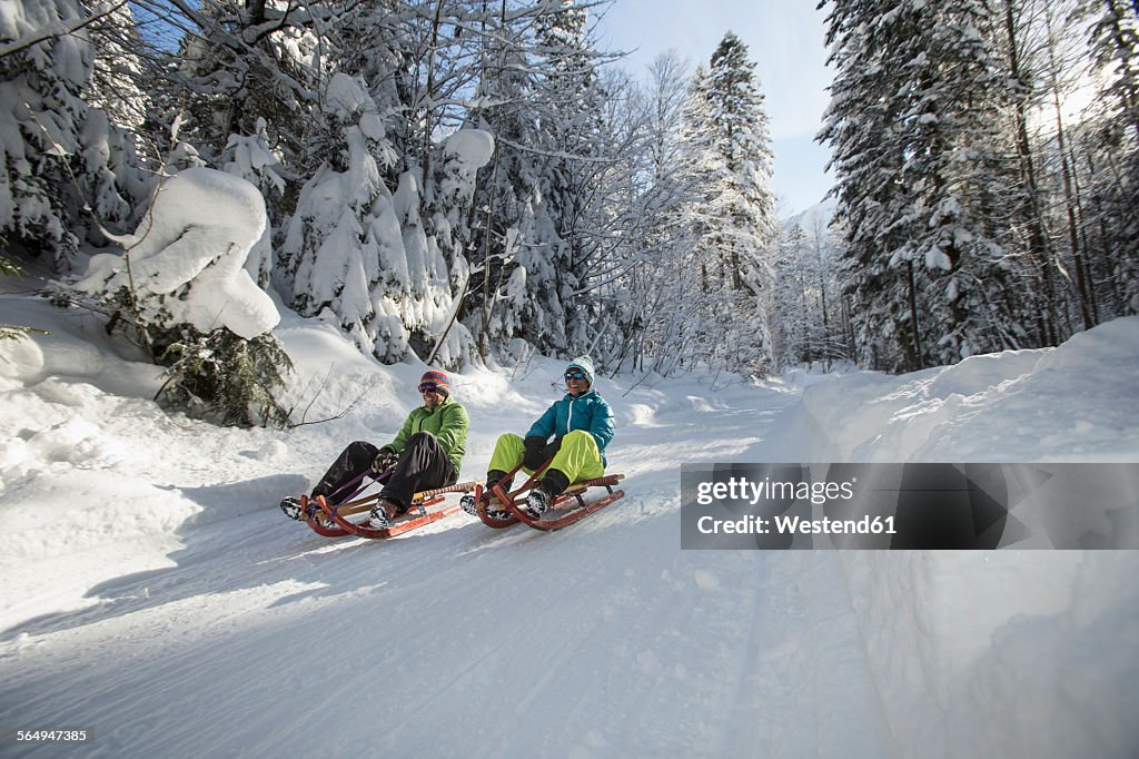 Germany, Bavaria, Inzell, couple having fun on sledges in snow-covered landscape