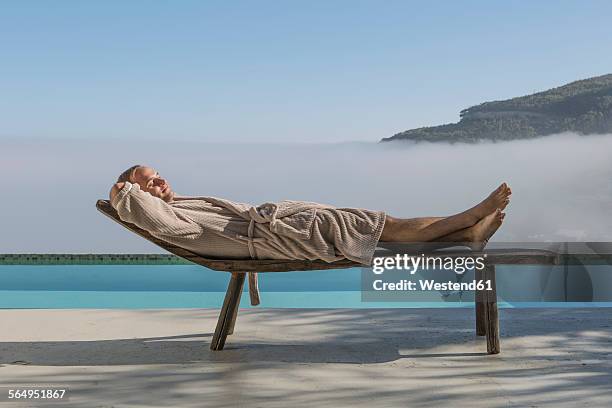 man in bathrobe lying on a lounge next to pool - bademantel stock-fotos und bilder