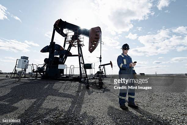 a man in overalls and hard hat at a pump jack in open ground at an oil extraction site. - mining hats stock pictures, royalty-free photos & images