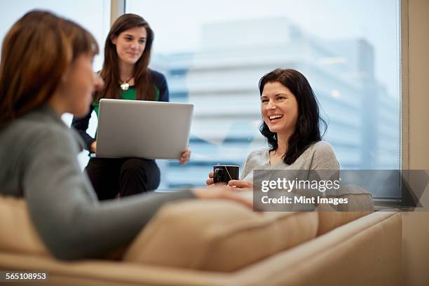three businesswomen by a window in an office, one holding a laptop. - editorial office stock pictures, royalty-free photos & images