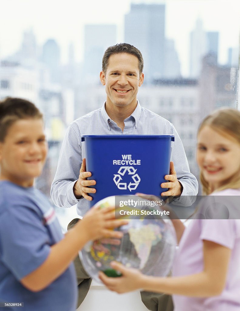 Portrait of a father holding a garbage bin with his two children standing in front of him