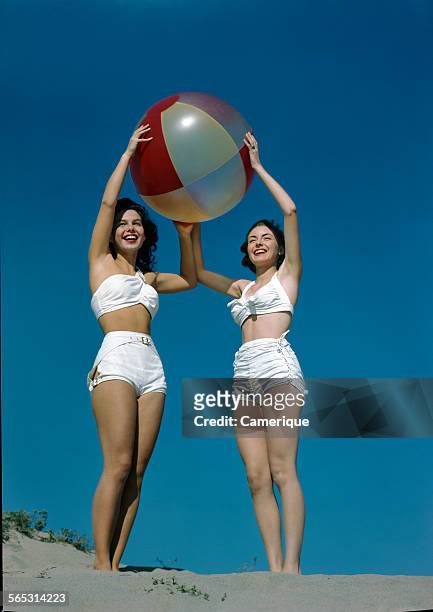 Portrait of two young girls in white bathing suits holding a large beach ball, Los Angeles, California, 1950.