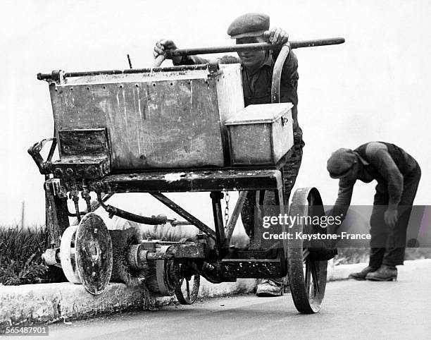 Un homme poussant un outil sur roulettes pour appliquer la peinture blanche de façon continue sur les bords de la route, un autre fait les finitions...