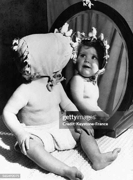 Assis devant une glace, bébé semble admirer son bonnet pascal orné de fleurs de printemps et de petits poussins, en France le 3 mars 1956.