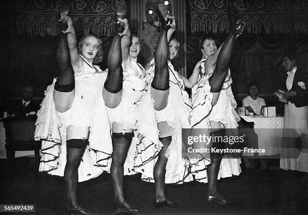 Quatre danseuses de cancan lèvent haut la jambe en porte-jarretelles et en se tenant par le talon sous le regard de spectateurs et d'un serveur.