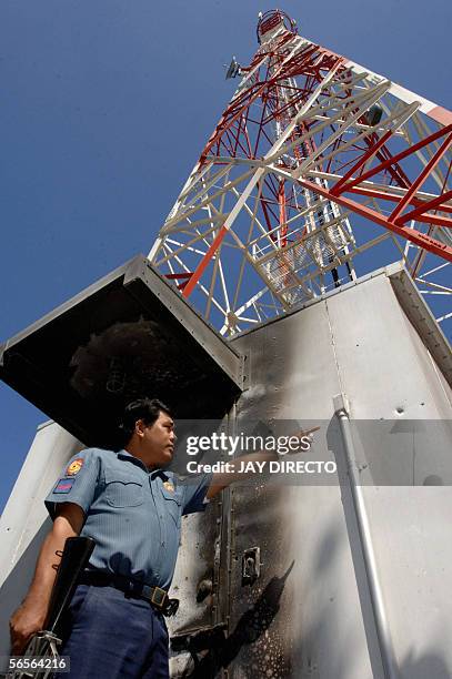 Police officer armed with an assault rifle points to bullet holes that tore through the partly burned metal enclosure housing transmitter equipment...