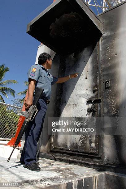 Police officer armed with an assault rifle points to bullet holes that tore through the partly burned metal enclosure housing transmitter equipment...