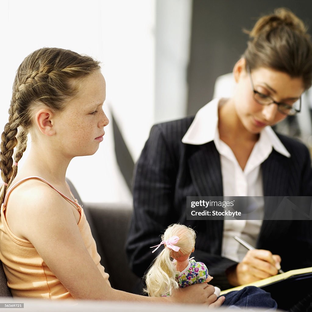 Female psychiatrist writing on a note pad and a girl playing with a doll beside her