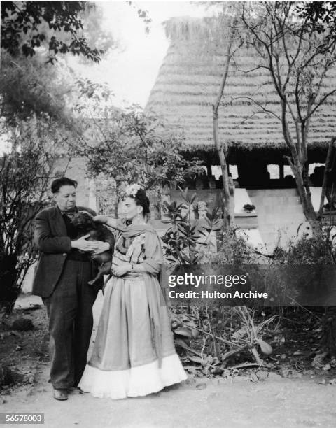 Married Mexican painters Frida Kahlo and Diego Rivera stand together with a pet dog in front of thatchted-roof hut which houses a number of...