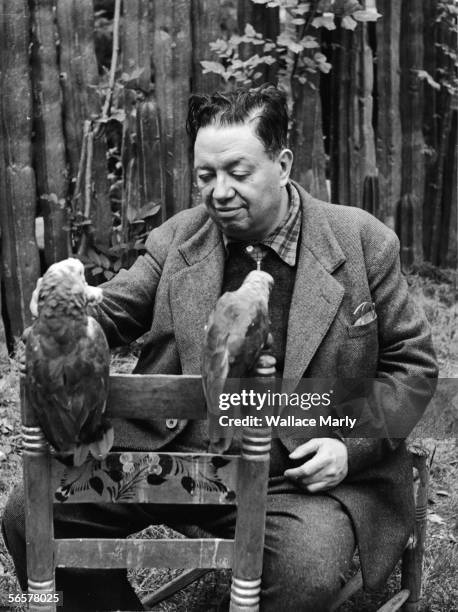 Mexican painter Diego Rivera sits outside and feeds a pair of parrots perched on the back of a painted chair, 1940s.