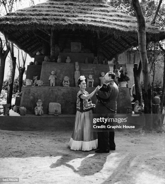 Married Mexican painters Frida Kahlo and Diego Rivera stand together with a pet monkey in front of thatchted-roof hut which houses a number of...