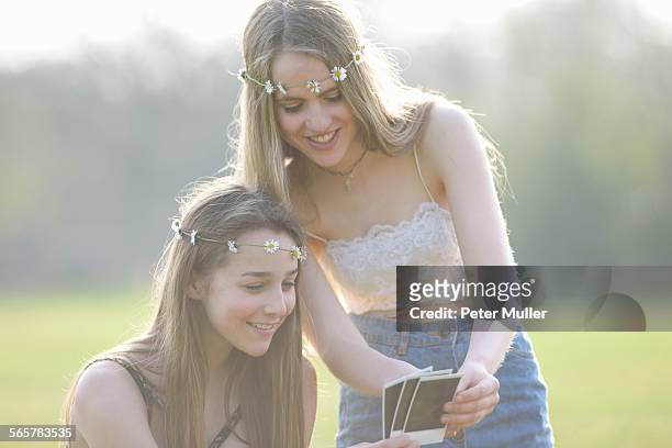 two teenage girls wearing daisy chain headdresses looking at instant photographs in park - ketting van madeliefjes stockfoto's en -beelden