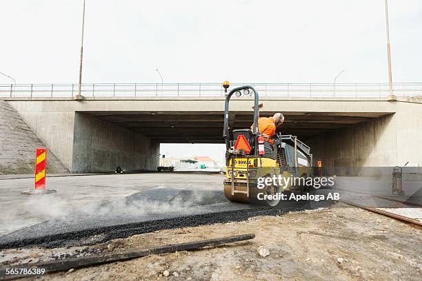 manual worker driving steamroller on hot tar at road construction site - wals stockfoto's en -beelden