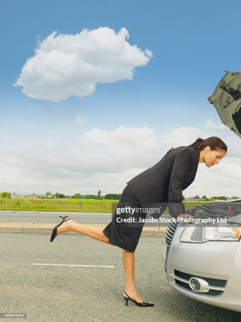 Caucasian businesswoman examining broken-down car engine on rural road
