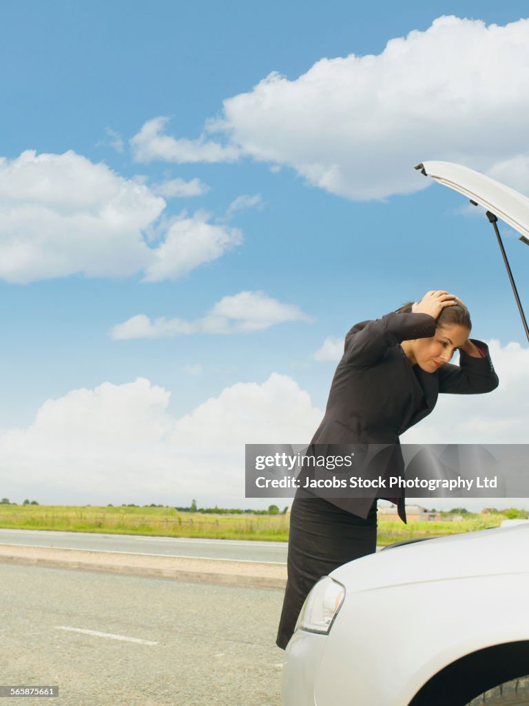 Caucasian businesswoman examining broken-down car engine on rural road