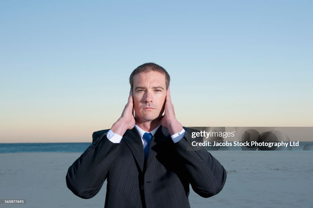 Caucasian businessman covering ears on beach