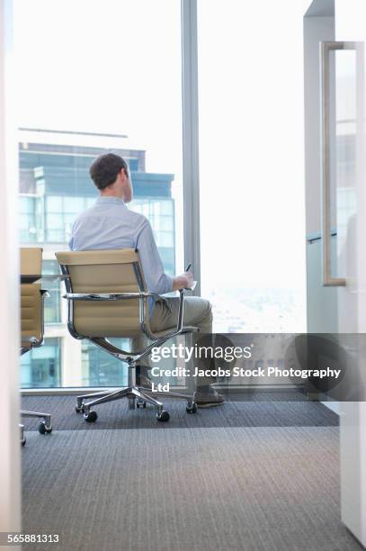 caucasian businessman looking out office window - office chair back fotografías e imágenes de stock