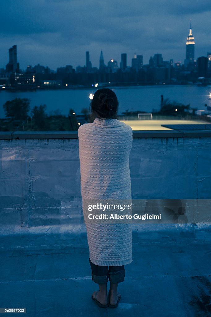 Caucasian woman wrapped in blanket admiring urban waterfront from rooftop