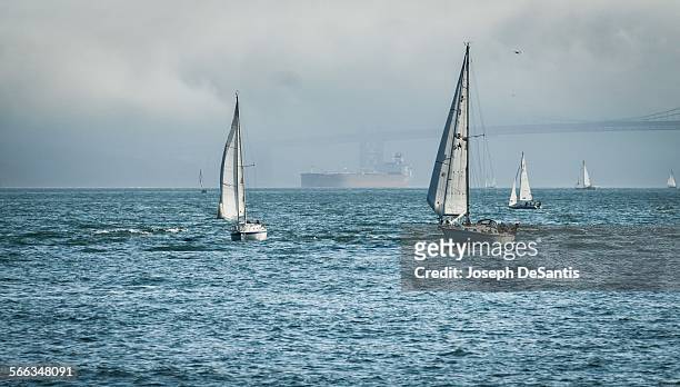 Seven sailboats in the foreground, and an oil tanker and the Golden Gate Bridge in the background, on a cloudy day.