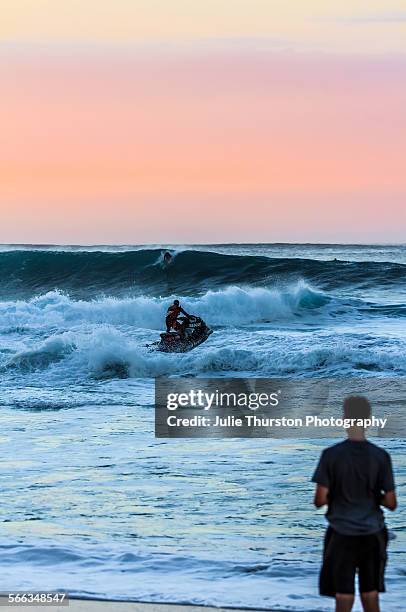 Rescue jet ski heads out as surfers on surfboards ride huge waves under a peaceful peach orange sky at Sunset Beach a famous tourist travel...