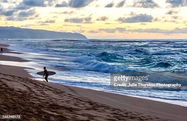 Surfer carrying surfboard comes in after surfing large waves at Sunset Beach famous for big surf and golden sunsets. Located on the North Shore of...