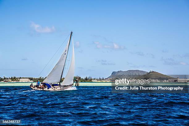 White sailboat with wind blowing through the sails, floats in the teal blue waters of Kaneohe Bay with the lush Kaneohe Marine Corps Base peninsula...