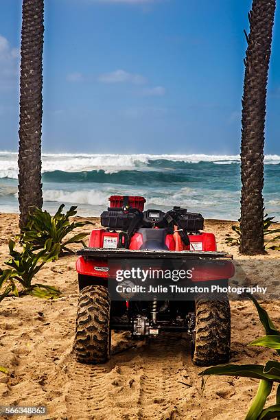Surfs up by a lifeguard red ATV beach buggy and fins facing the teal blue choppy water on a white sandy palm tree lined beach under blue skies at the...