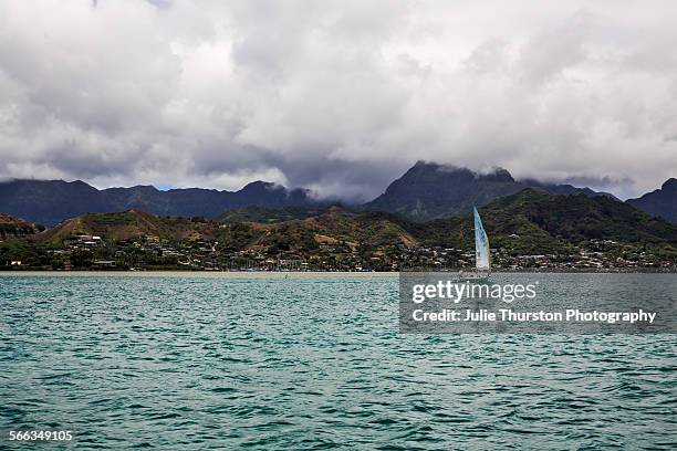 White sailboat with wind blowing through the sails, floats in the waters of Kaneohe Bay with the beautiful Ko'olau Mountain Range and lush shoreline...