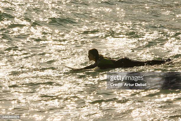 Girl laying on surf board rowing with hands and the sun reflected on the Mediterranean Sea in the Barcelona shoreline. Vilassar de Mar, Catalonia,...