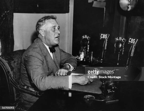 American President Franklin Delano Roosevelt sits at a desk in front of a series of microphones as he delivers a 'Fireside Chat' radio broadcast,...