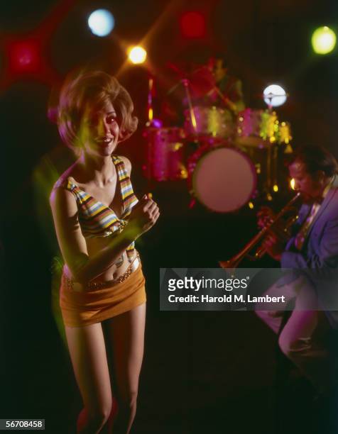Woman wearing a mini skirt dancing in a night club, a man is playing the trumpet behind her, 1969.