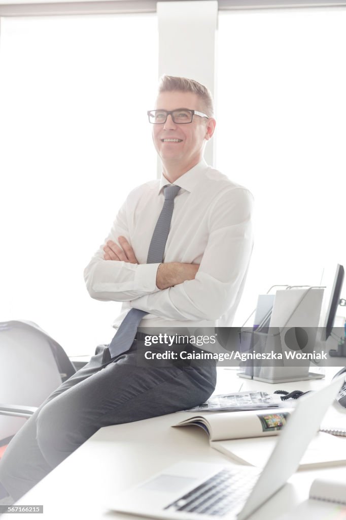 Portrait confident businessman with eyeglasses sitting on desk with arms crossed