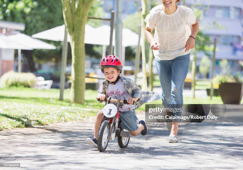 Mother chasing son riding bicycle with helmet in sunny park