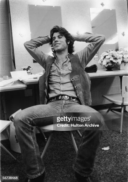 American actor John Travolta sits in a chair and leans back with his hands behind his head, late 1970s.