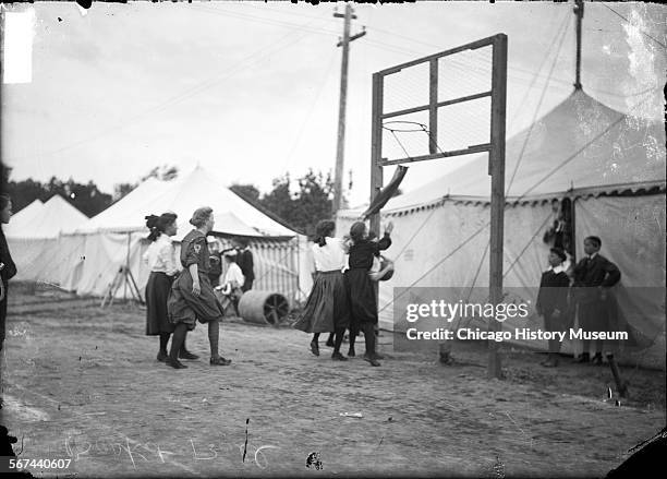 Girls playing basketball on a grass court surrounded by light colored tents, Chicago, Illinois, 1905. A group of boys is standing near the basket,...