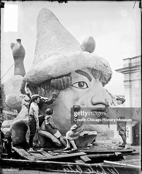 Portrait of two boys and two girls standing and sitting in front of a giant model head of a man wearing a hat at the White City amusement park,...